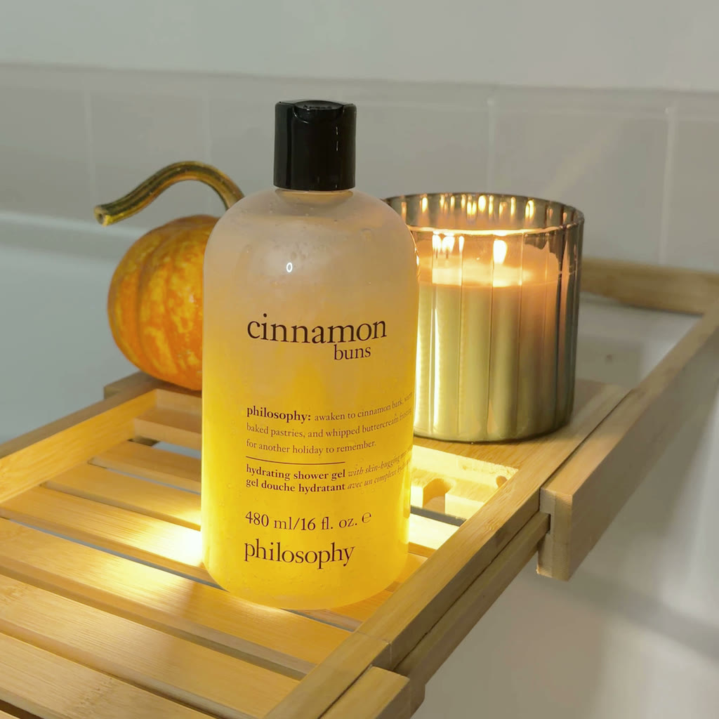 philosophy Cinnamon Buns bubble bath on a bamboo bath tray beside a lit candle and a small pumpkin